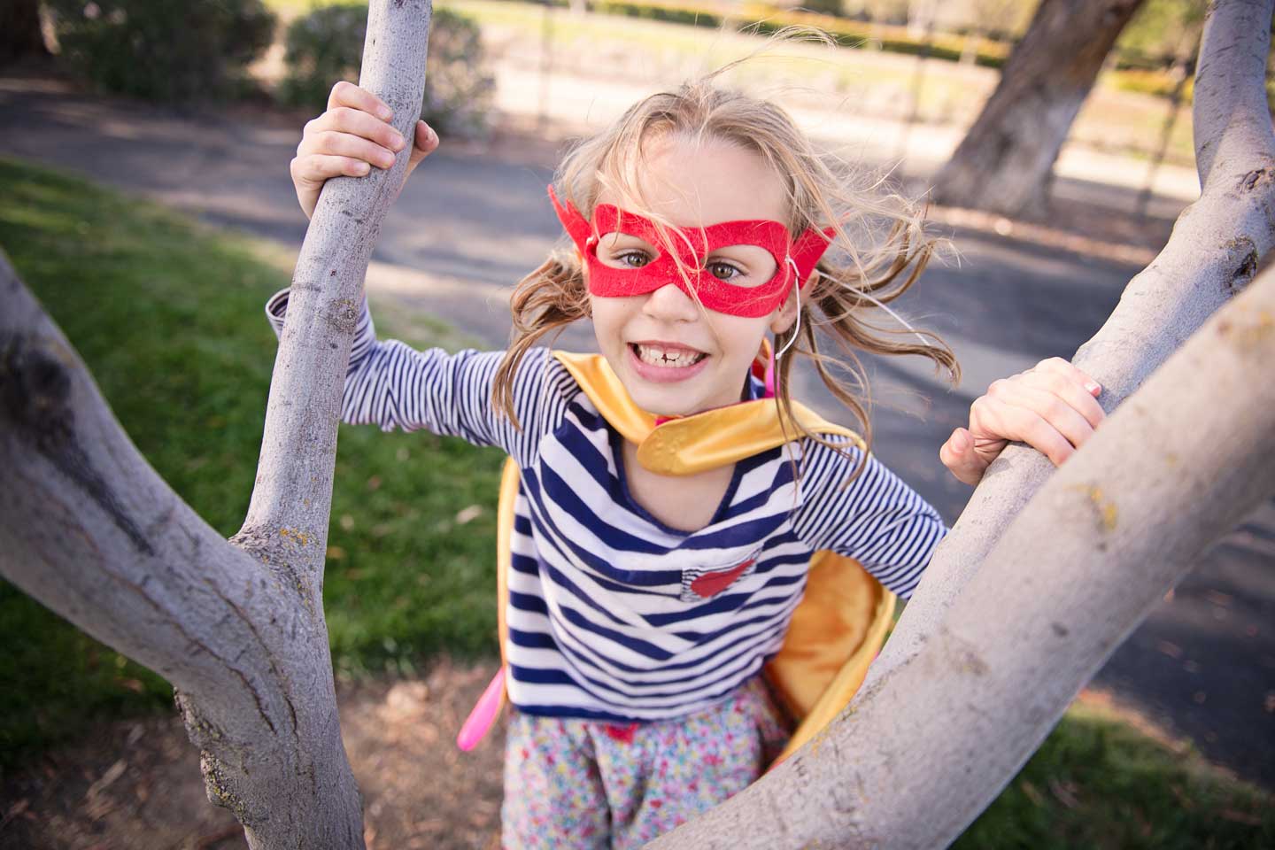 Ein kleines Mädchen mit Cape und Superheldenmaske klettert in einem Baum.